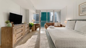 A modern hotel room with a large bed, a TV, a wooden dresser, a sofa, and a view of tall buildings. - ICON W BRICKELL RENTALS SLEEK STUDIO #2005 (Miami)