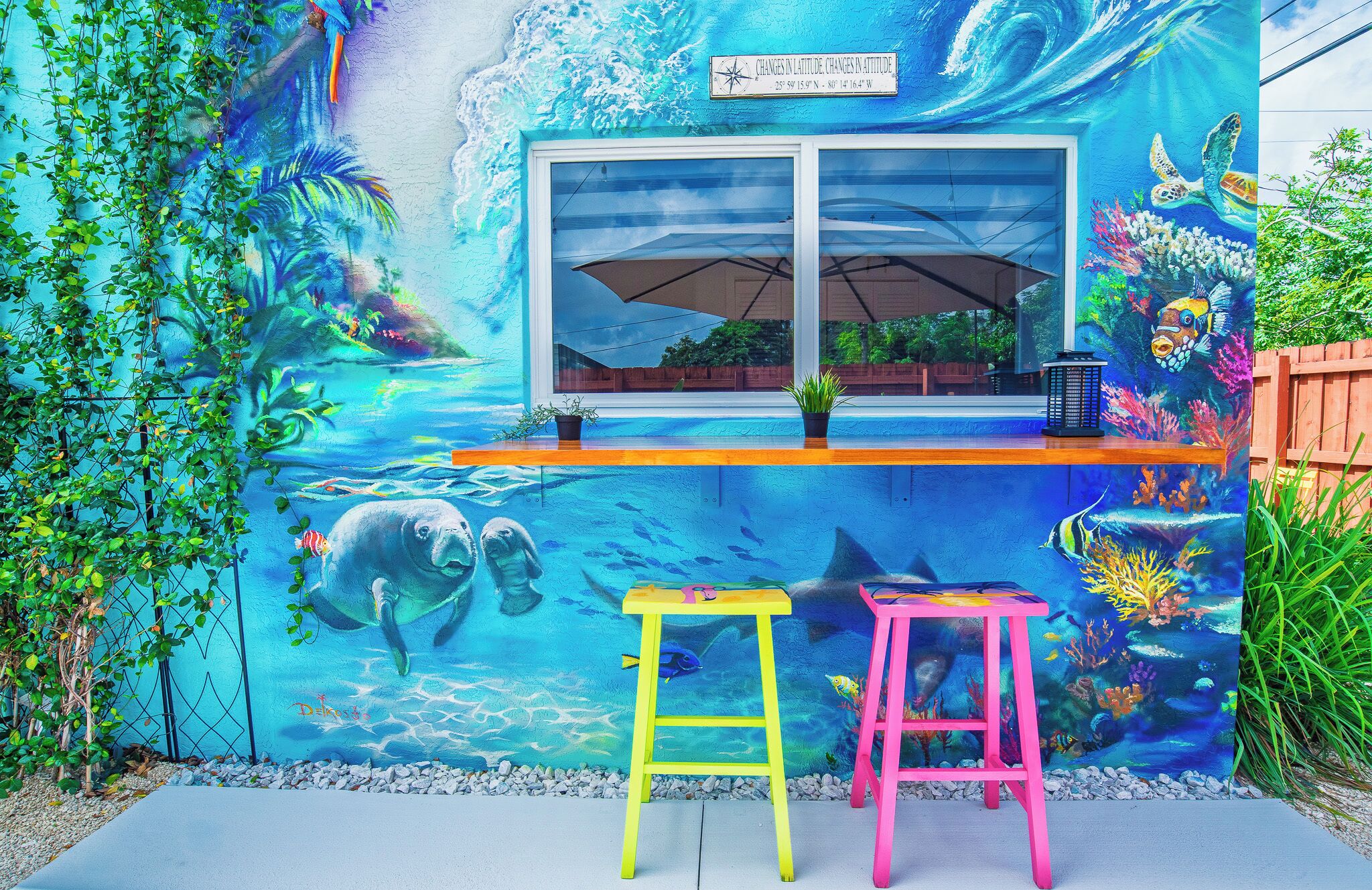 Un mural vibrant amb una finestra, dos tamborets colorits i una taula petita amb una planta.