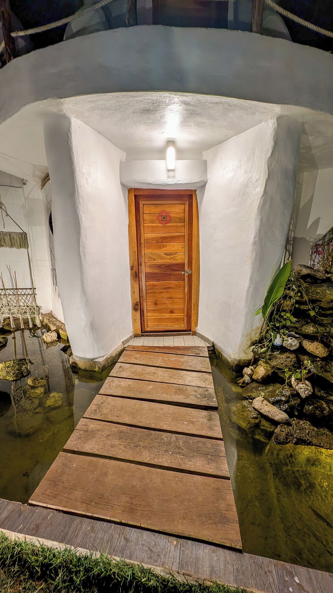 A wooden door with a light fixture above it, leading to a white-walled room with a small wooden dock extending into a pond.