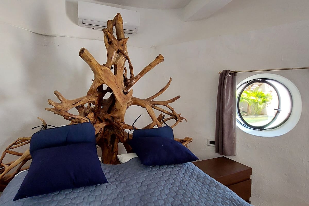 Ein Schlafzimmer mit einem großen Baumstamm als Mittelpunkt, einem Rundfenster und einem Bett mit blauer Bettwäsche.