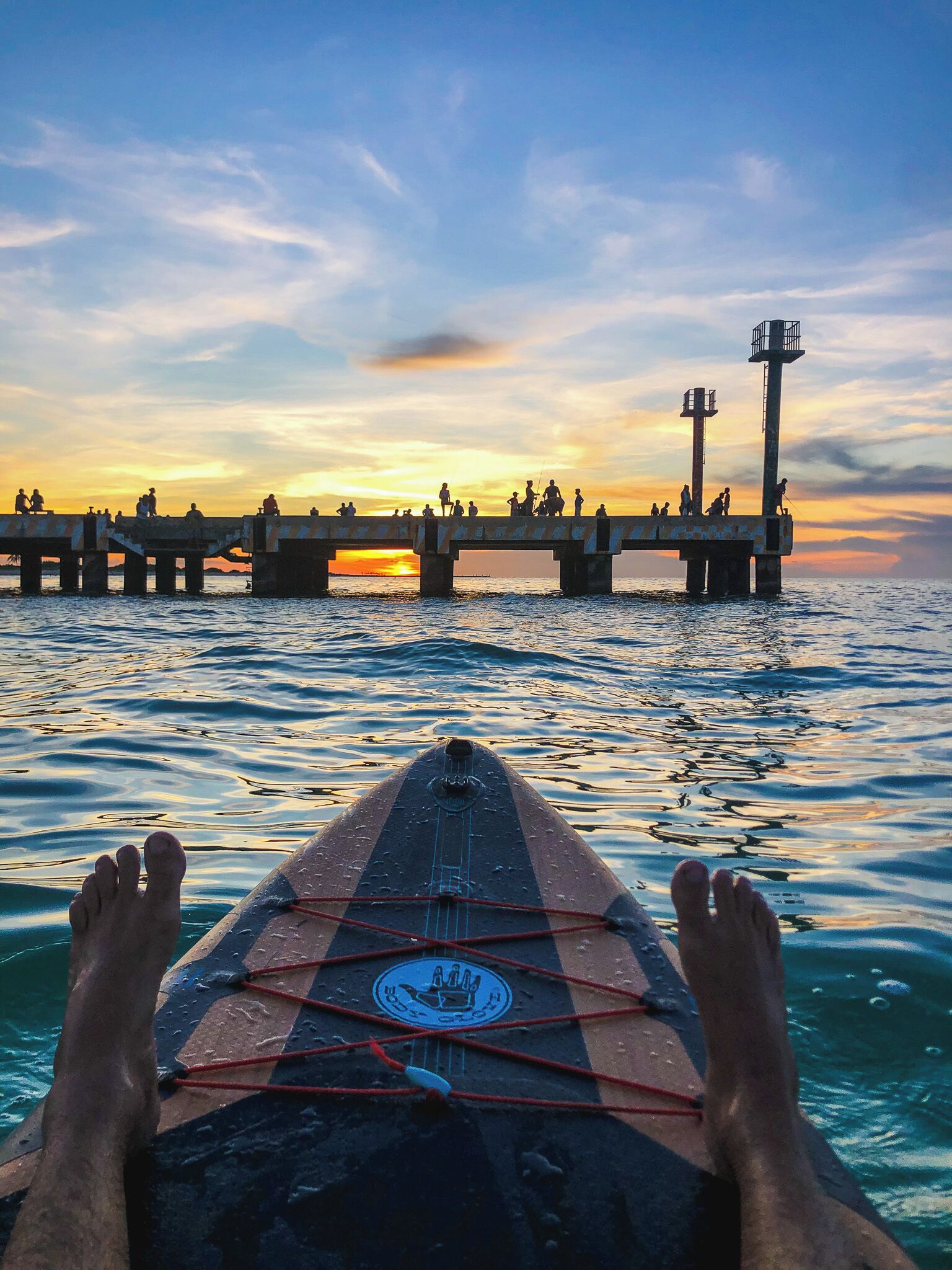 Iemand ligt op een paddleboard, kijkend naar een pier waar mensen op lopen, tijdens zonsondergang.
