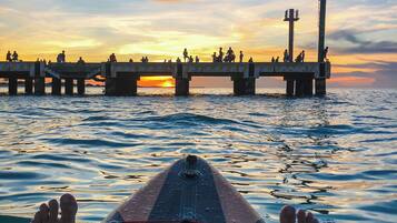 Iemand ligt op een paddleboard, kijkend naar een pier waar mensen op lopen, tijdens zonsondergang.