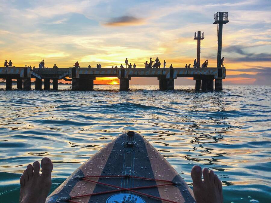Iemand ligt op een paddleboard, kijkend naar een pier waar mensen op lopen, tijdens zonsondergang.