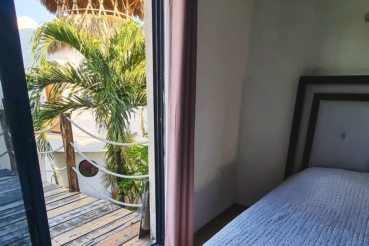 Ein Schlafzimmer mit einem großen Bett, einem Fenster mit Blick auf ein Reetdachhaus und Palmen sowie einem Balkon mit Holzboden.