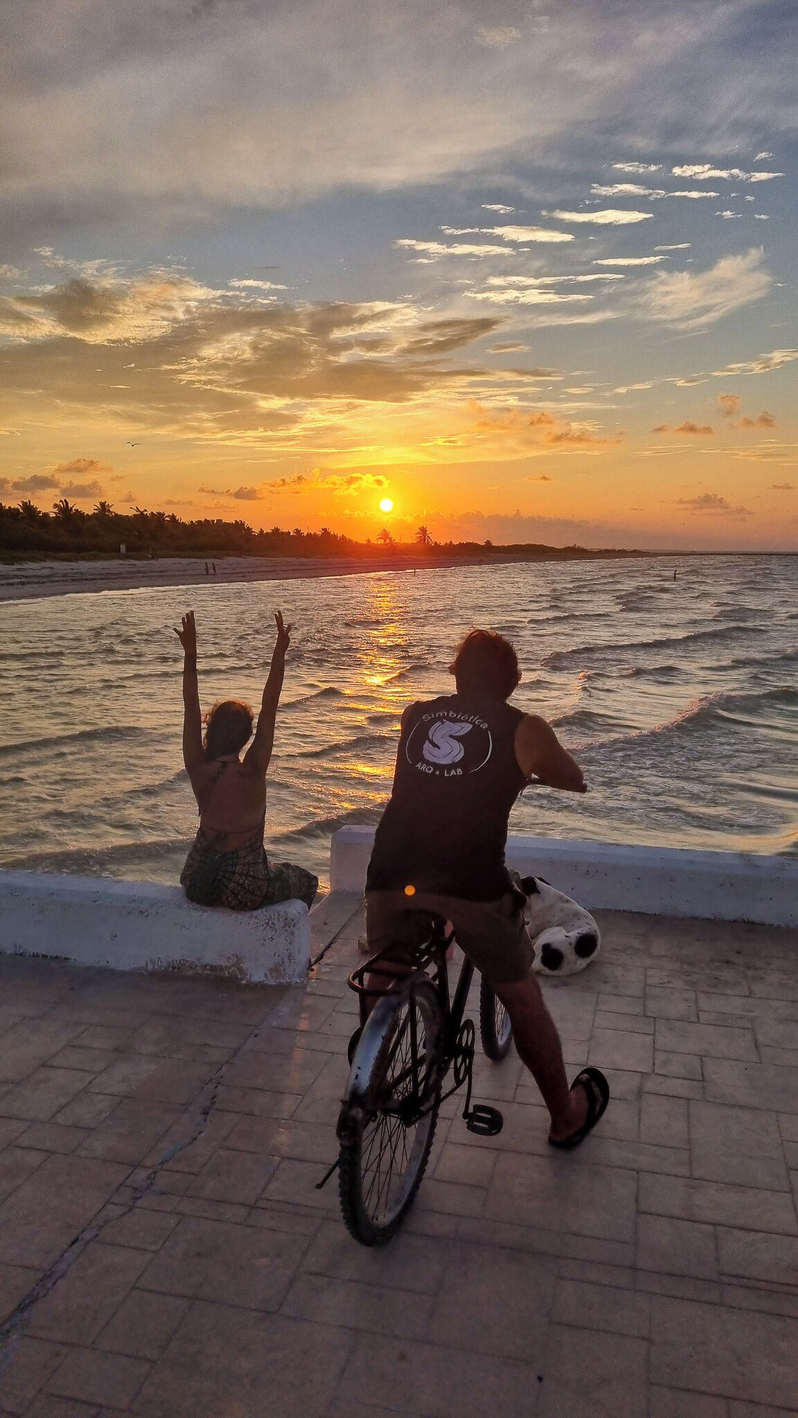 Deux personnes se trouvent au bord de la mer au coucher du soleil : l’une est assise et l’autre est debout, tenant un vélo.