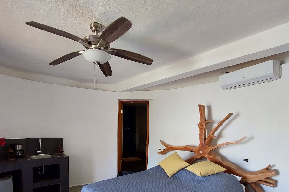 Ein Schlafzimmer mit einem Bett, einem Deckenventilator, einem Holz-Kopfteil, einem Schreibtisch mit Waschbecken und einer Tür zu einem anderen Raum.
