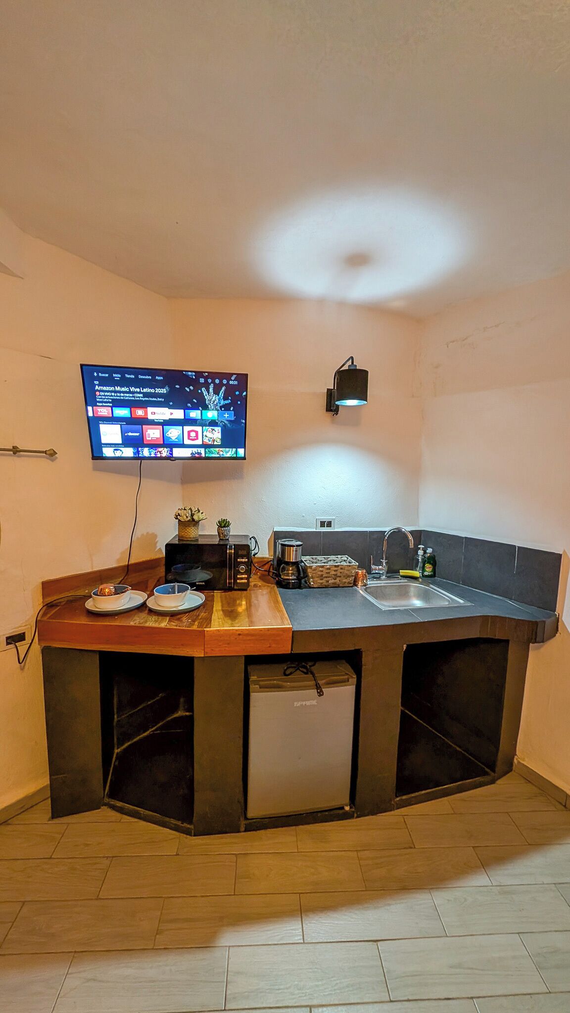 A compact kitchen with a built-in microwave, a sink, and a small refrigerator. A flat-screen TV is mounted on the wall displaying a music streaming service.