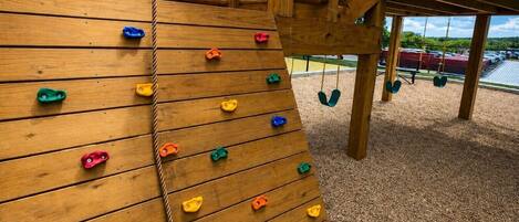 Un mur d’escalade en bois, doté de différentes prises de couleurs variées, une corde et un ensemble de balançoires en arrière-plan.