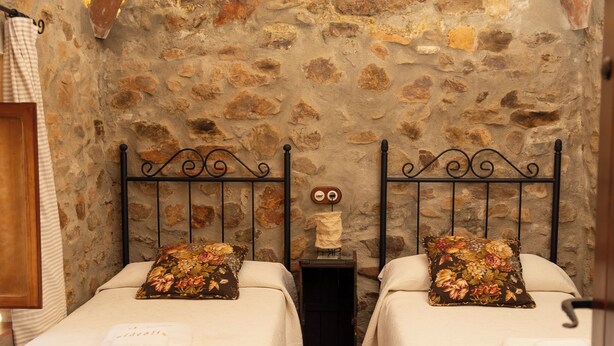 Una stanza rustica e accogliente con due letti, ognuno con un cuscino fiorito e una coperta bianca. Le pareti sono in pietra a vista, e si nota una porta in legno.