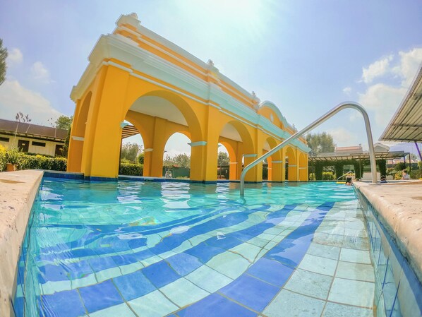 Une piscine au sol carrelé, avec un bâtiment jaune orné d’arches en arrière-plan.