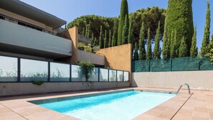 A modern pool area with a clear blue pool, tiled deck, and a view of a building with a balcony and tall trees in the background.