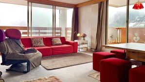 A spacious living area with a red sofa, a small table with a lamp, and a view of a snowy landscape through large windows. - Courchevel Moriond - Apartment 6 ppl, 3 bdr, Private Parking (Courchevel)