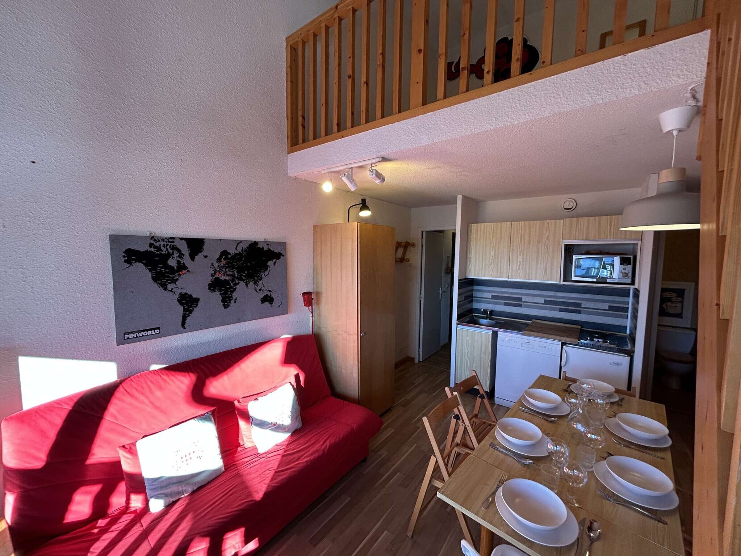 A compact living space with a red sofa, a dining table set for four, a kitchen area with a microwave, and a world map on the wall.