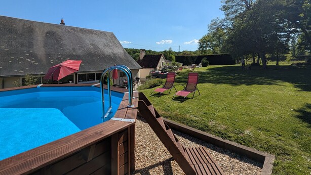 Una piscina con plataforma de madera, un edificio de piedra y una zona verde con sillas.
