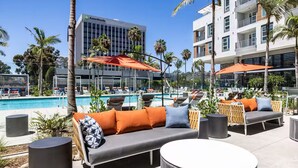 A poolside lounge area with outdoor seating, a pool, and a view of a hotel building.