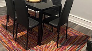 A dining area with a black table and chairs, a colorful rug, and a framed elephant artwork on the wall.