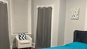 A bedroom with a bed, a chair, a window with curtains, and a wall-mounted artwork.