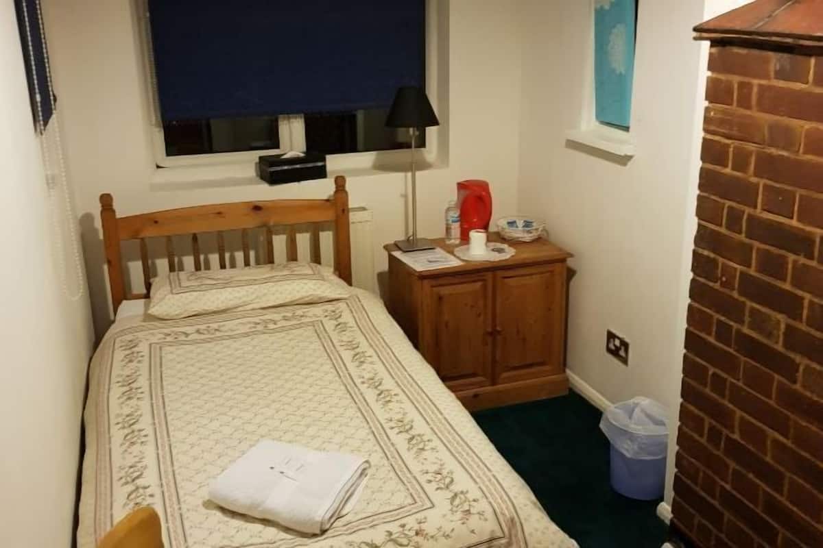 A small, neatly arranged bedroom with a single bed, a wooden cabinet, a window with a blind, and a brick wall.