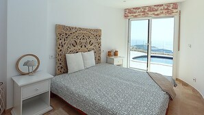 Un dormitorio con una cama grande, una mesita de noche, un aparador y vistas a una piscina a través de un ventanal corredera.