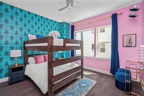 A bunk bed with a white and pink bedding, a nightstand with a lamp, a blue patterned wall, a large window with blue curtains, and a mirror with lights.