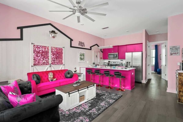 A modern living room with a pink sofa, black armchair, and a coffee table. A kitchen with pink cabinets and a bar area is visible in the background.
