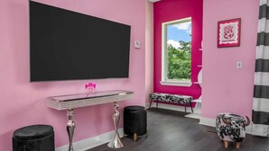 A room with a large flat-screen TV mounted on a pink wall, a small table with a decorative top, and a bench with a black and white patterned cover.