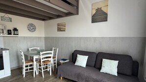 A modern living room with a grey sofa, white chairs, a small round table, and a kitchen area with a fridge and cabinets.