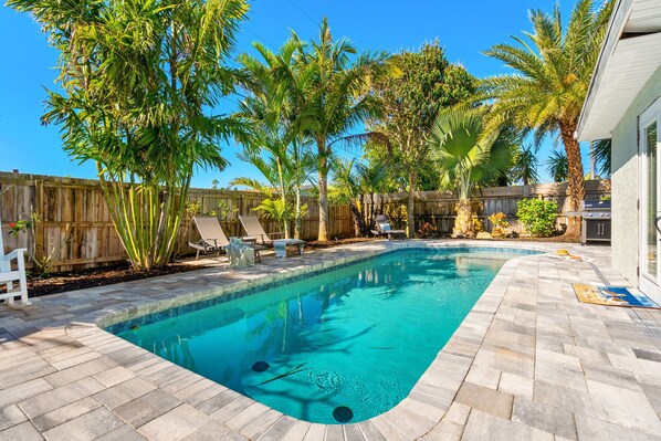 Une piscine rectangulaire, bordée d’une terrasse carrelée, entourée de palmiers et d’une clôture en bois.