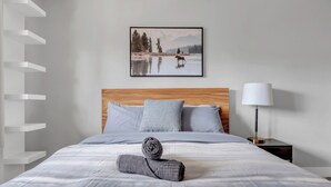 A neatly made bed with a rolled towel, a framed picture of a moose by a lake, and a bedside lamp.