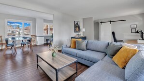 A modern living room with a grey sofa, wooden coffee table, and a dining area with a white table and chairs.