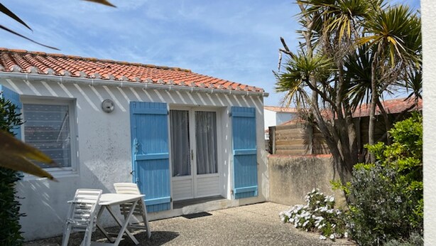 Una casa pequeña con techo de tejas, fachada blanca y contraventanas azules. Se ve una silla blanca y una mesa pequeña en el exterior.