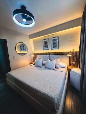 A modern bedroom with a bed, bedside lamps, a wall-mounted mirror, and framed artwork.