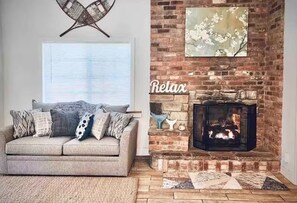 A cozy living room with a fireplace, a sofa with patterned cushions, and a window with blinds. - Entire House with private lake access and hot tub! Near local ski resorts!!  (Honeoye)