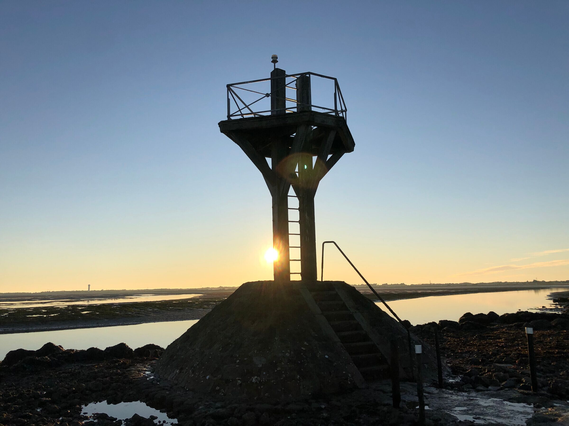 A tower structure with a ladder leading up to a platform, surrounded by a rocky landscape and a body of water.
