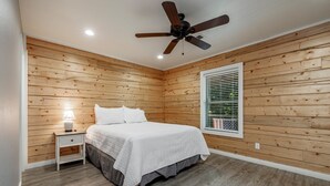 Ein Schlafzimmer mit Holzwandverkleidung, einem Bett mit weißer Bettwäsche, einem Nachttisch mit Lampe, einem Deckenventilator und einem Fenster mit Jalousien.