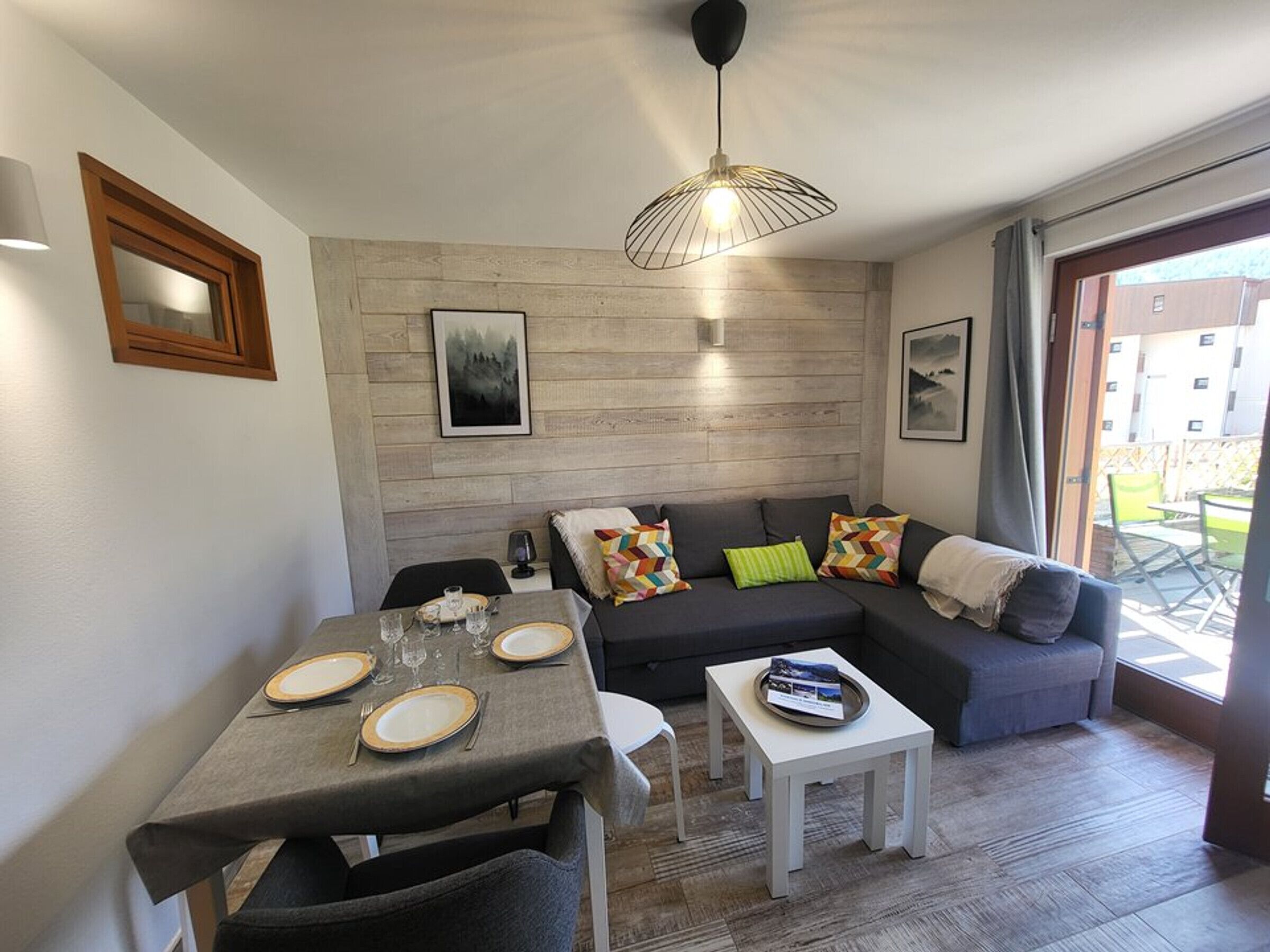 A modern living room with a dining area, grey sofas, colorful cushions, a wooden wall, and a large window.