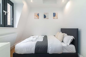 Un dormitorio con cama, ropa de cama blanca, un radiador, una ventana con persianas y tres obras de arte colgadas en la pared.
