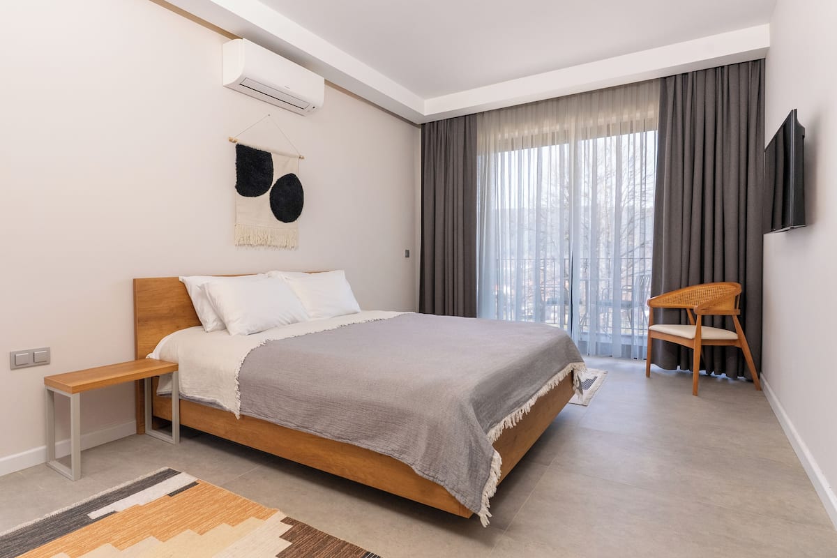 Quarto moderno com cama de madeira, mesa de cabeceira, cadeira e uma janela grande com cortinas.
