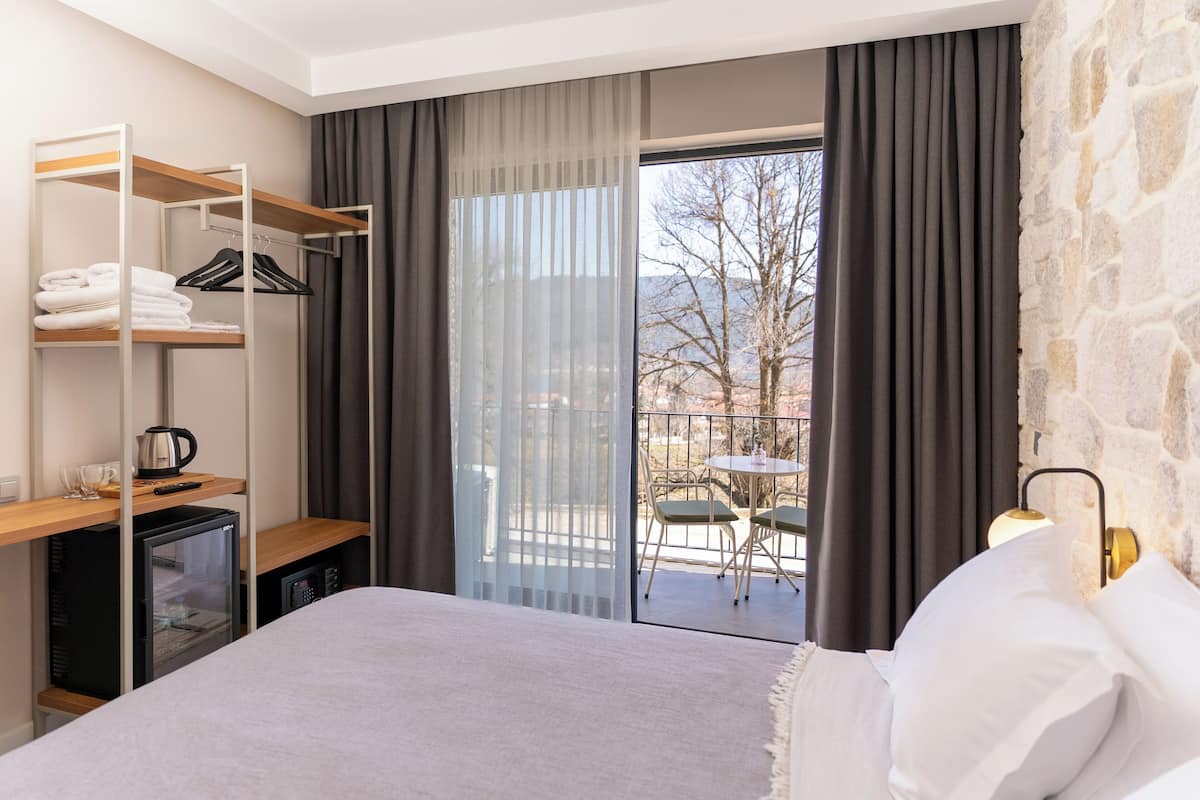 Quarto de hotel com cama, guarda-roupa, mini-frigorífico, máquina de café e varanda com mesa e cadeiras.