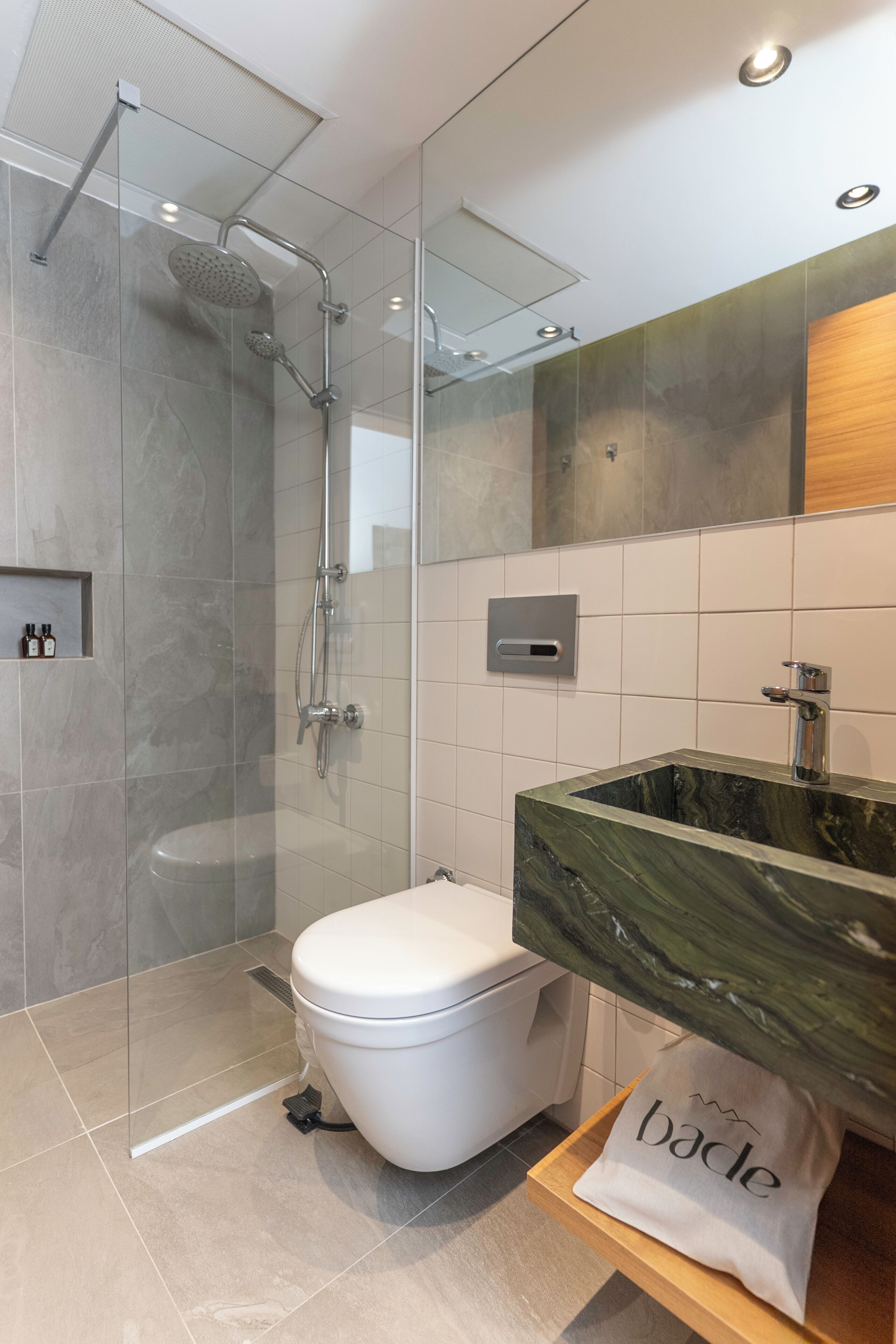 Un ba ño moderno con ducha de vidrio, inodoro blanco, lavamanos de mármol y repisa de madera con una bolsa etiquetada "bade".
