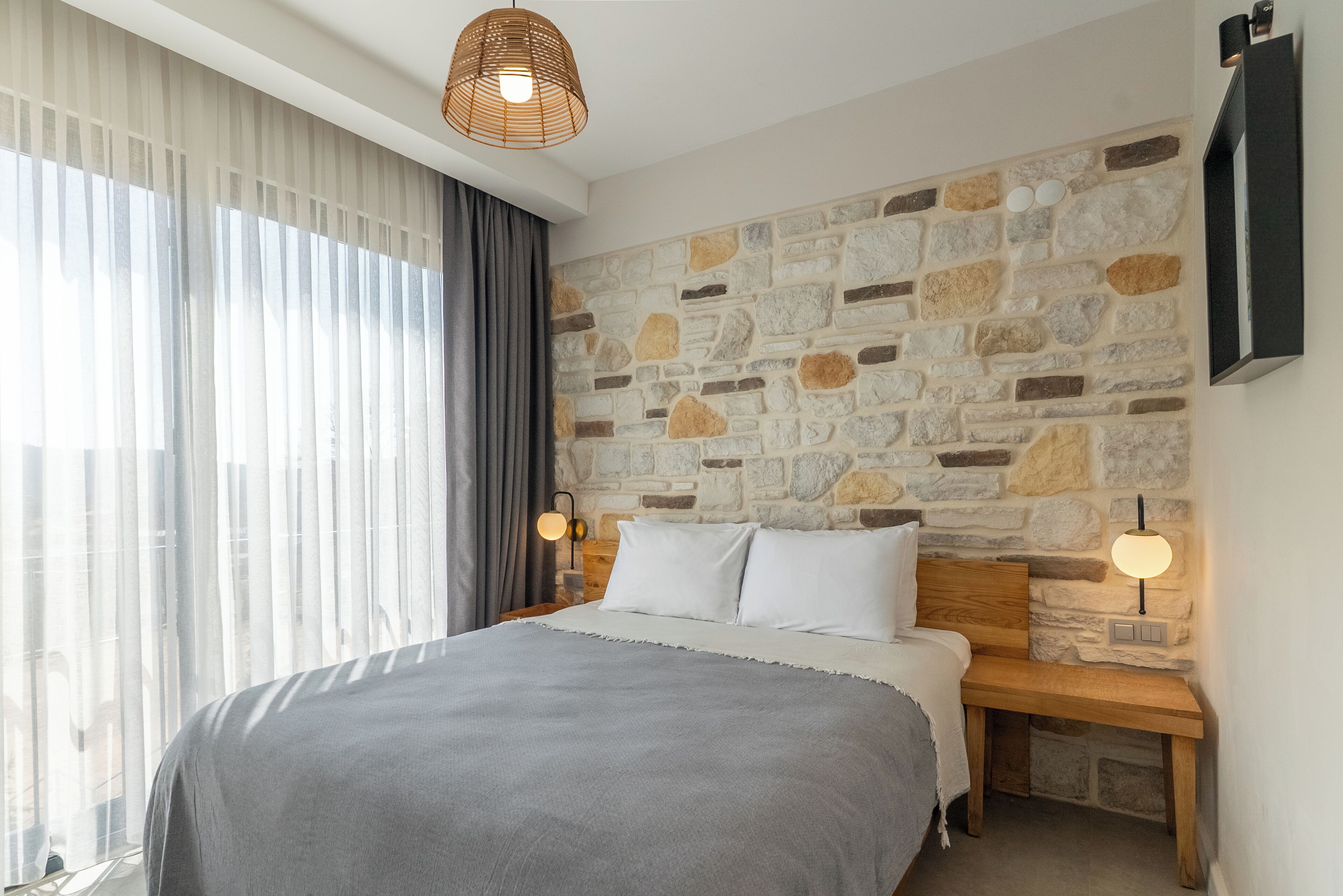 Un dormitorio con pared de piedra, una cama con manta gris, mesitas de noche de madera y dos lámparas fijadas a la pared.