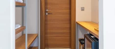 Un pasillo estrecho con una puerta de madera, una estantería metálica con ropa doblada y una pequeña mesa con una silla.