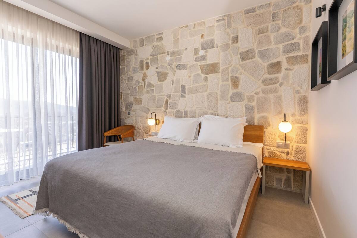 Um quarto com parede de pedra, uma cama grande, duas mesas de cabeceira com abajures, uma janela com cortinas e vista para a cidade.