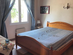 Um quarto com cama de madeira, janela com cortinas, mesa de cabeceira com abajur e um quadro na parede.
