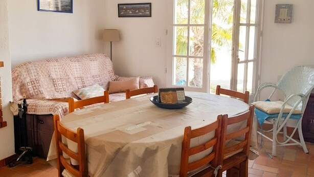 A cozy room with a round wooden table, a sofa, and a chair. There is a window with a view of palm trees outside. - Semi-detached house 4 pers, near beach, pets allowed (La Guérinière)