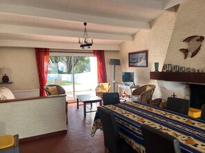 A spacious living area with a large sofa, a coffee table, and a dining table with a patterned tablecloth. There is a chandelier hanging from the ceiling, a television on a stand, and a fireplace with decorative items on the mantel. - Comfortable house 300 m from the beach, pets allowed (La Guérinière)