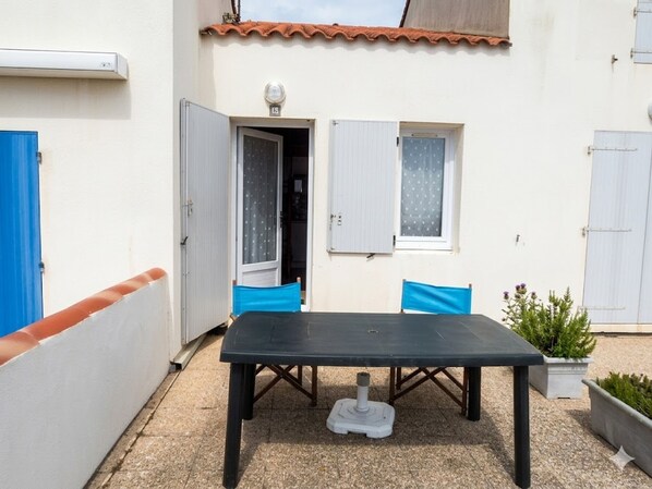 An outdoor patio area with a table and chairs, a small window, and a door leading to a room.
