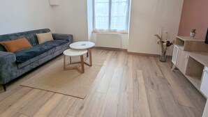 Un salón con un sofá azul oscuro, dos mesas redondas blancas, piso de madera, una ventana con persianas, un reloj de pared y una planta pequeña en un estante.