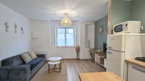Un salon moderne comprenant un canapé, une petite table ronde, une table à manger en bois, un coin cuisine avec un réfrigérateur et un four à micro-ondes, et une fenêtre avec des rideaux.