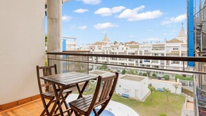 Een balkon met een houten tafel en stoelen, met uitzicht op een zwembad en een woongebied met witte gebouwen en een blauwe lucht.